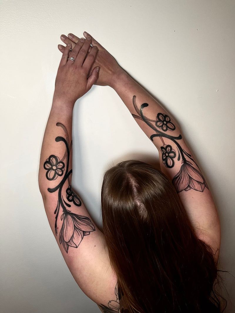 A person with long brown hair seen from behind, stretching both arms overhead with black floral tattoo designs visible on both forearms against a plain off-white wall.