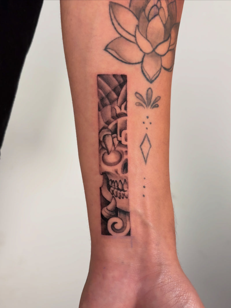 A close-up of a person's forearm featuring a black and gray tattoo design split into two vertical sections: one with a detailed skull and floral motif, and the other with simple geometric shapes and a lotus flower.
