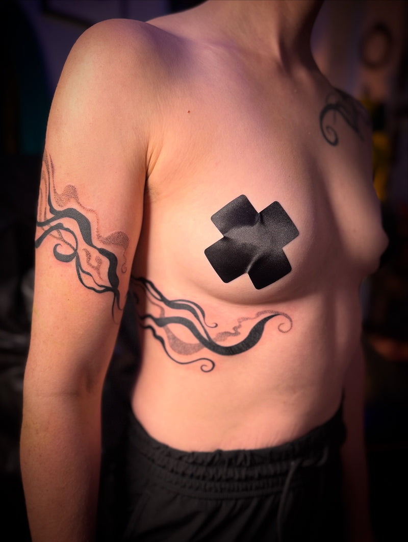 A close-up of a person's upper torso showing a black tattoo design on the chest and left arm, with a black cross-shaped bandage or patch applied to the chest. The person is wearing black shorts, and the background is dark and blurred.
