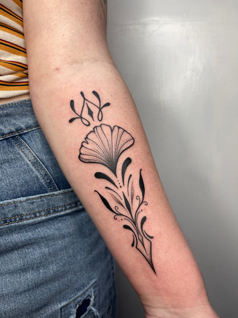 A close-up of a person's forearm featuring a black ink floral tattoo with stylized leaves and abstract flourishes above a fan-shaped flower. The person is wearing blue jeans and a striped top, standing against a plain light gray wall.