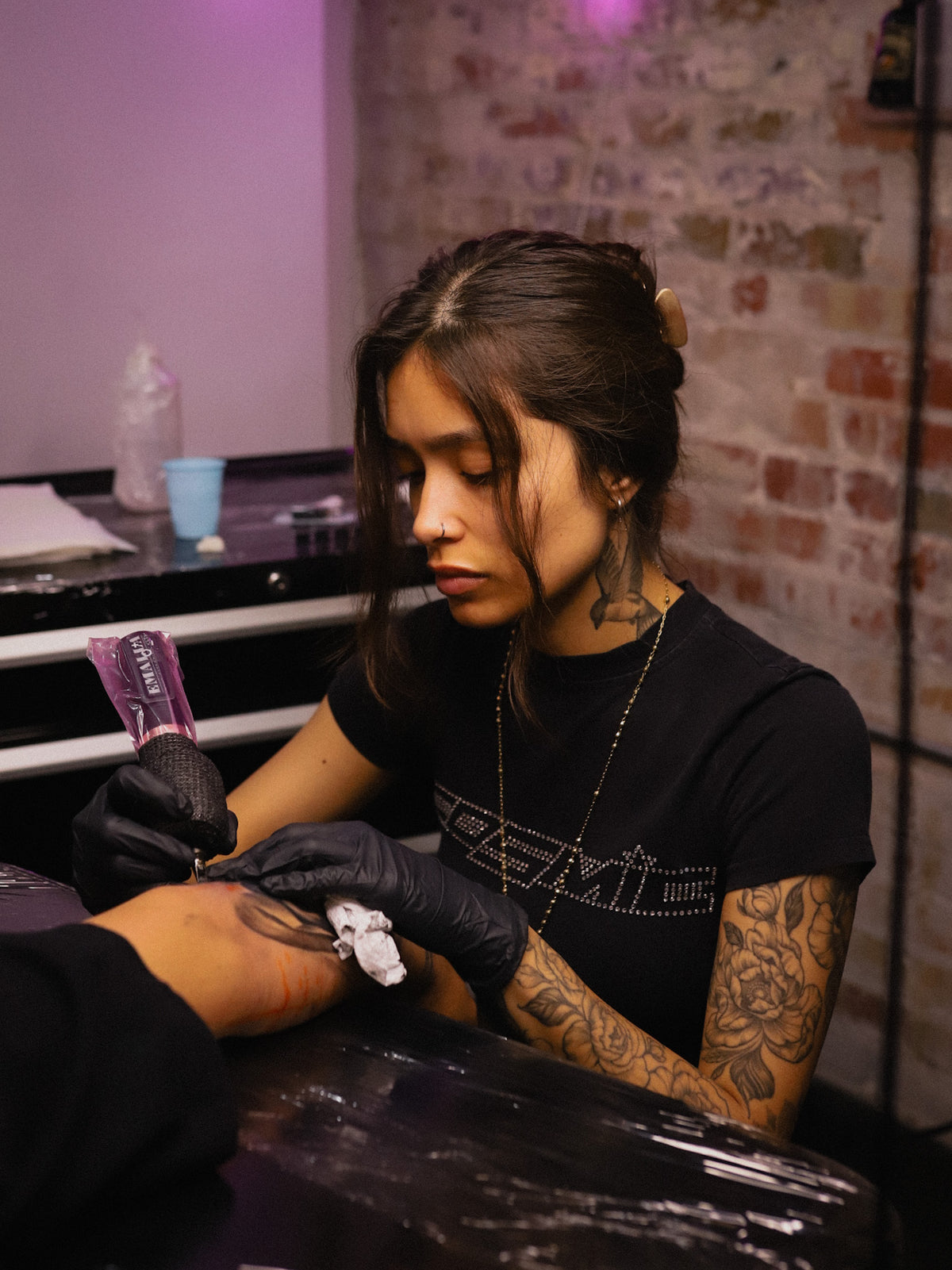 A tattoo artist wearing black gloves and a black t-shirt with a studded graphic, tattooing a client's arm in a tattoo studio with a brick wall background.