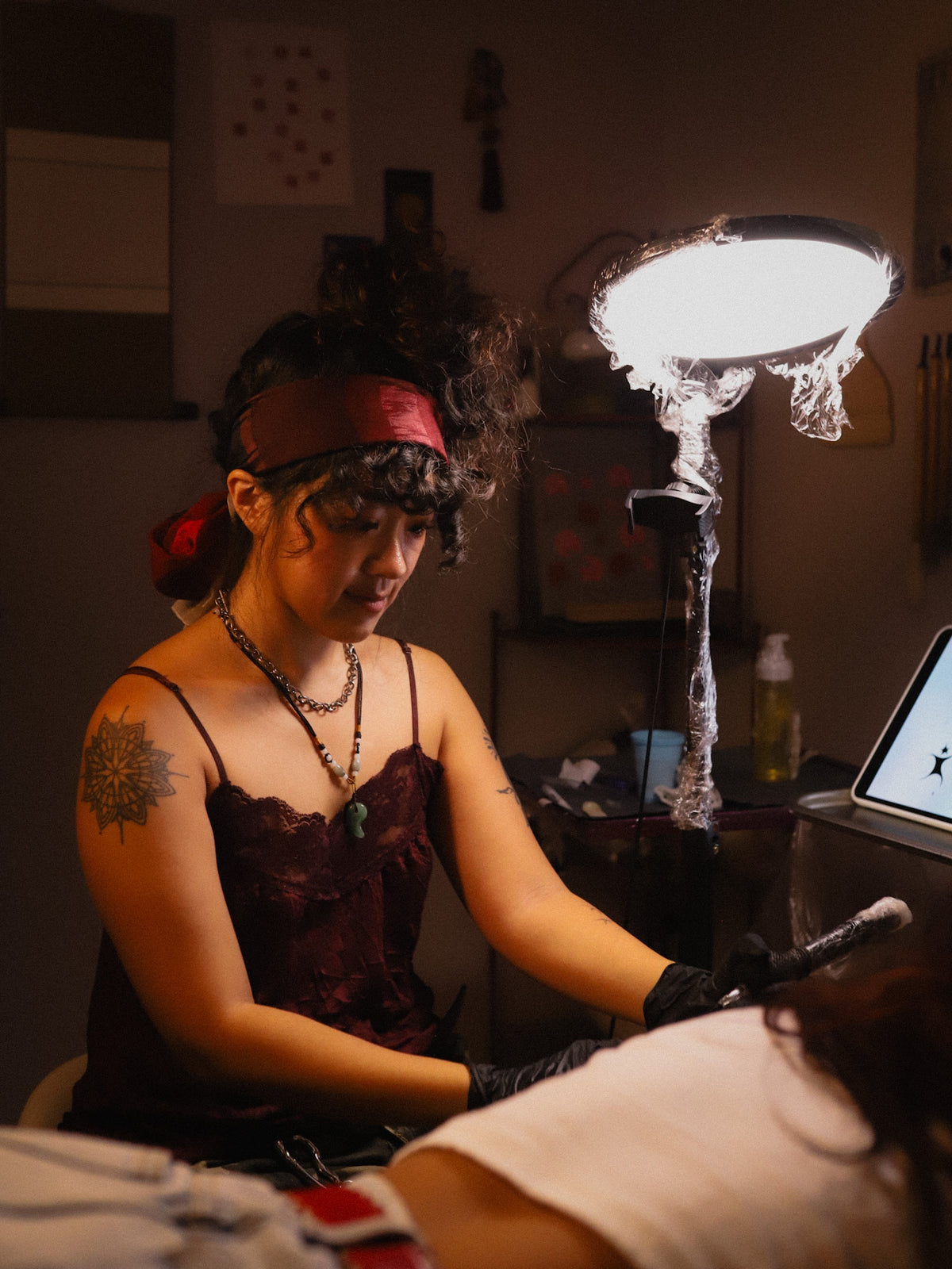 A tattoo artist wearing a dark red lace top and black gloves sits at a tattoo table under a bright ring light, with a person receiving a tattoo in the foreground.