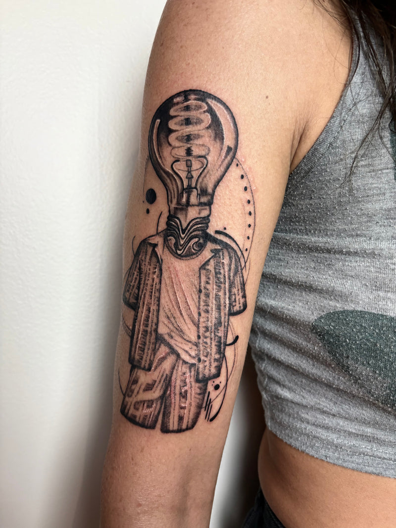 A close-up of a person's upper arm featuring a black and gray tattoo of a stylized figure with a light bulb for a head, wearing draped robes. The person is wearing a gray ribbed tank top against a plain off-white wall.
