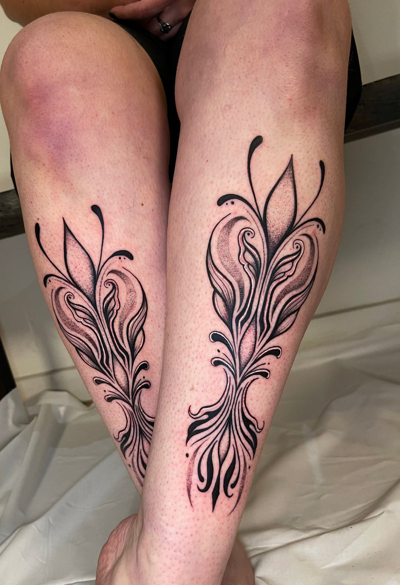 A close-up of a person's legs showing two matching black floral tattoo designs on the upper calves, with light skin and a white fabric surface in the background.