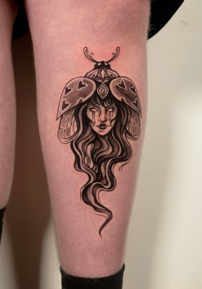 A close-up of a person's leg showing a detailed black and gray tattoo of a woman's face emerging from a moth-like insect with long flowing hair.