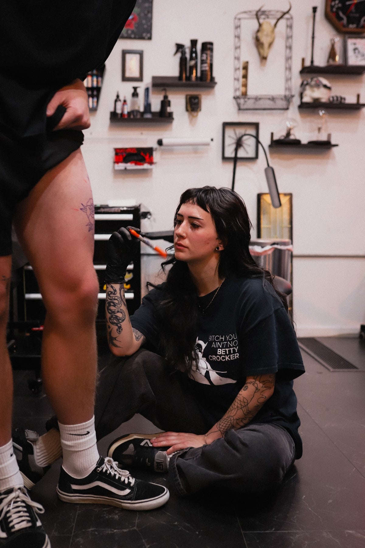 A tattoo artist sitting on the floor in a tattoo studio, wearing a black graphic t-shirt and dark gray pants, stenciling a client whose lower body is visible in the foreground.