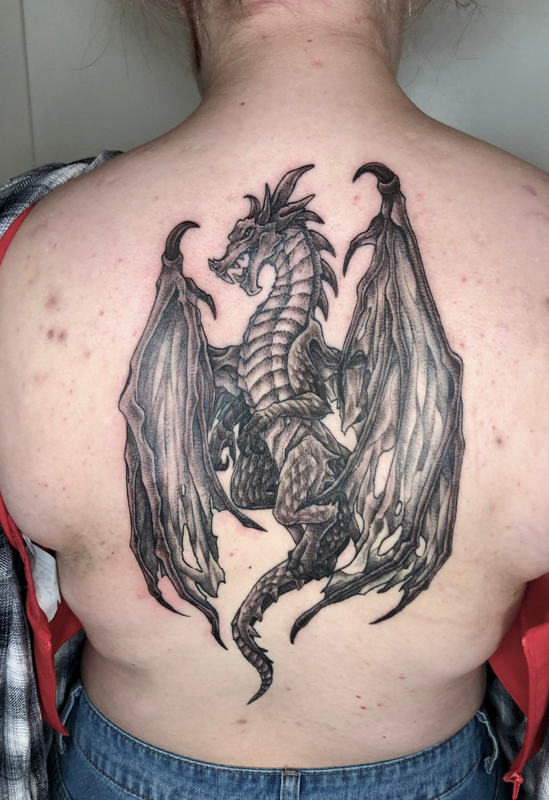 A large black and gray dragon tattoo on the back of a person's upper back, shown from behind. The tattoo features a detailed winged dragon in a grayscale ink style, with the person wearing a red shirt and blue jeans partially visible.