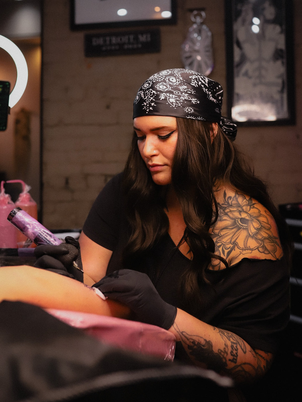 A tattoo artist wearing a black bandana, black shirt, and black gloves applies ink to a client's arm in a tattoo studio.