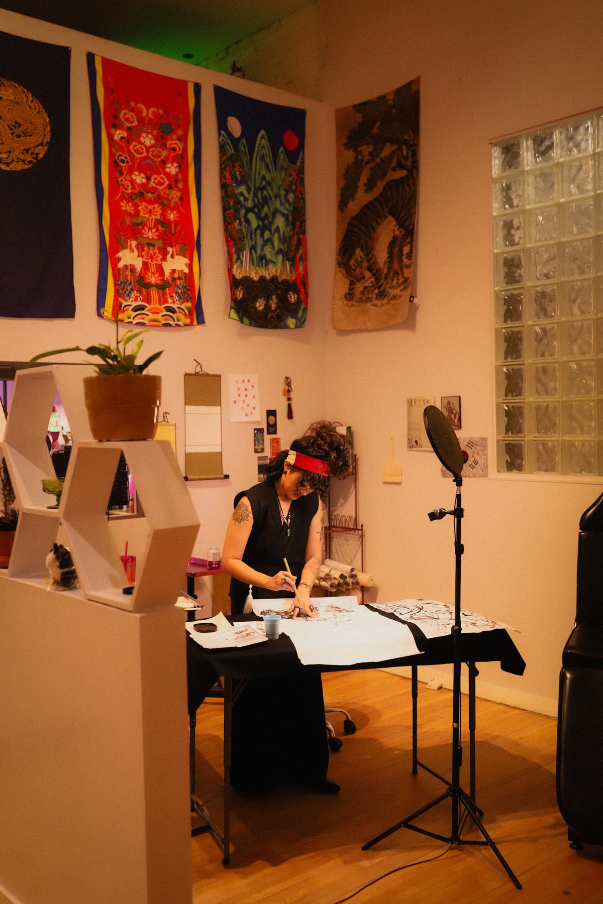 A person wearing a red headband sits at a table in a room, working on a piece of paper. The room has warm lighting, wooden floors, a glass block window, and several colorful hanging tapestries on the wall.