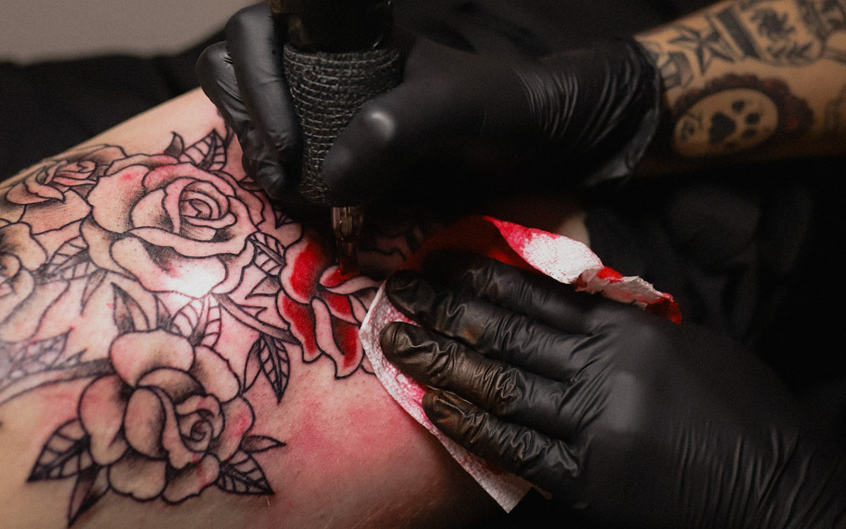A close-up of a tattoo artist's gloved hands tattooing a rose design onto a person's upper arm, with the black-and-gray floral tattoo and a red-and-white cleaning cloth visible.