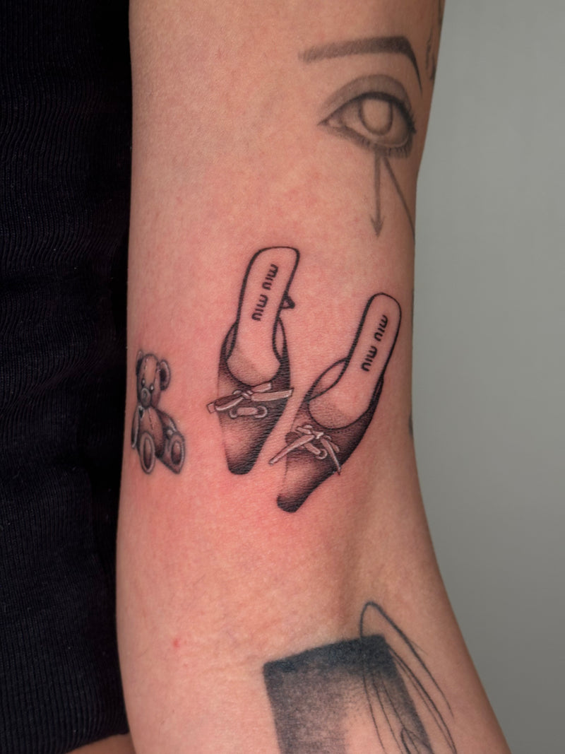 A close-up of a person's forearm featuring a black and gray tattoo of a pair of heels with 'miu miu' text.