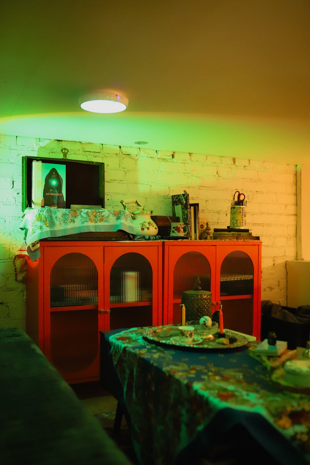 A red wooden cabinet with arched door panels stands against a white brick wall in a room with dramatic green and yellow ambient lighting. A patterned tablecloth-covered table is in the foreground, and framed items and decor sit on top of the cabinet.