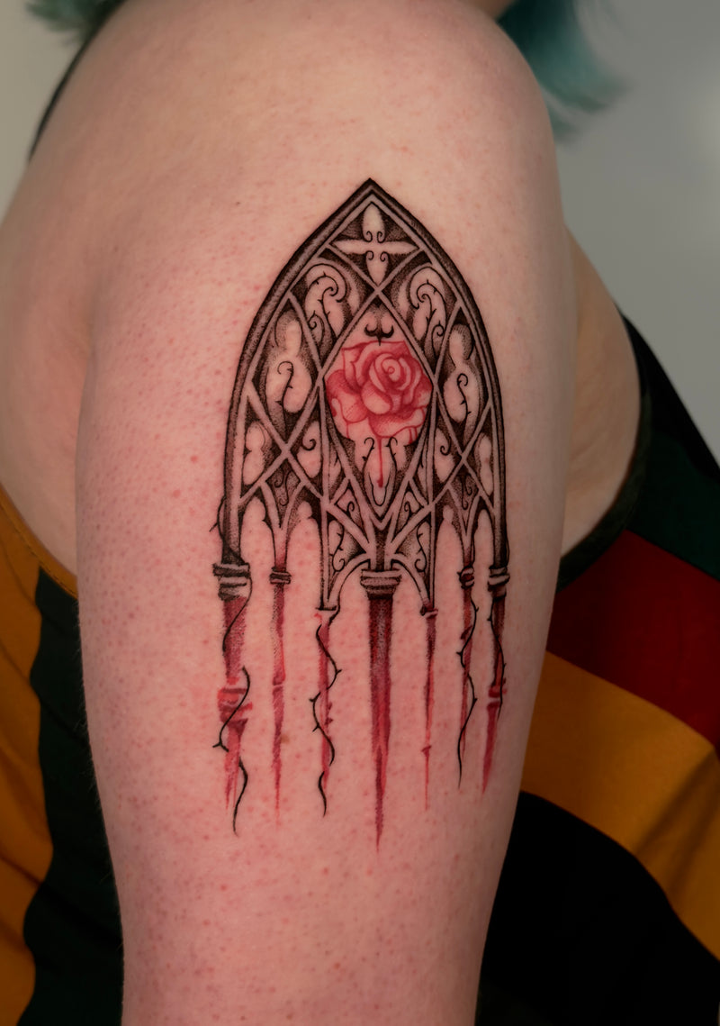 A close-up of a person's upper arm featuring a freshly tattooed gothic-style design. The black ink artwork depicts an ornate arched window frame with a central pink rose and dripping red accents, set against the wearer's skin.