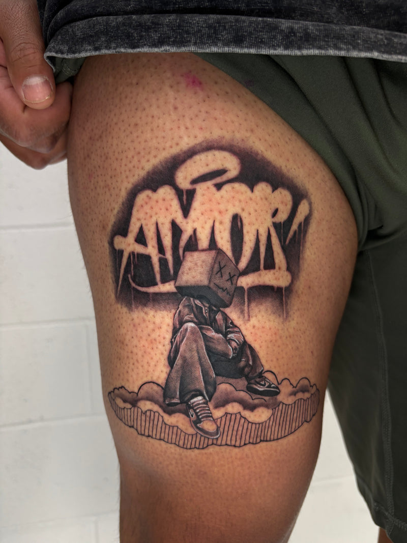 A close-up of a person's upper thigh showing a detailed black and gray temporary tattoo. The tattoo features a cartoon character with a box head sitting on clouds beneath graffiti-style text that reads 'AMOR'.