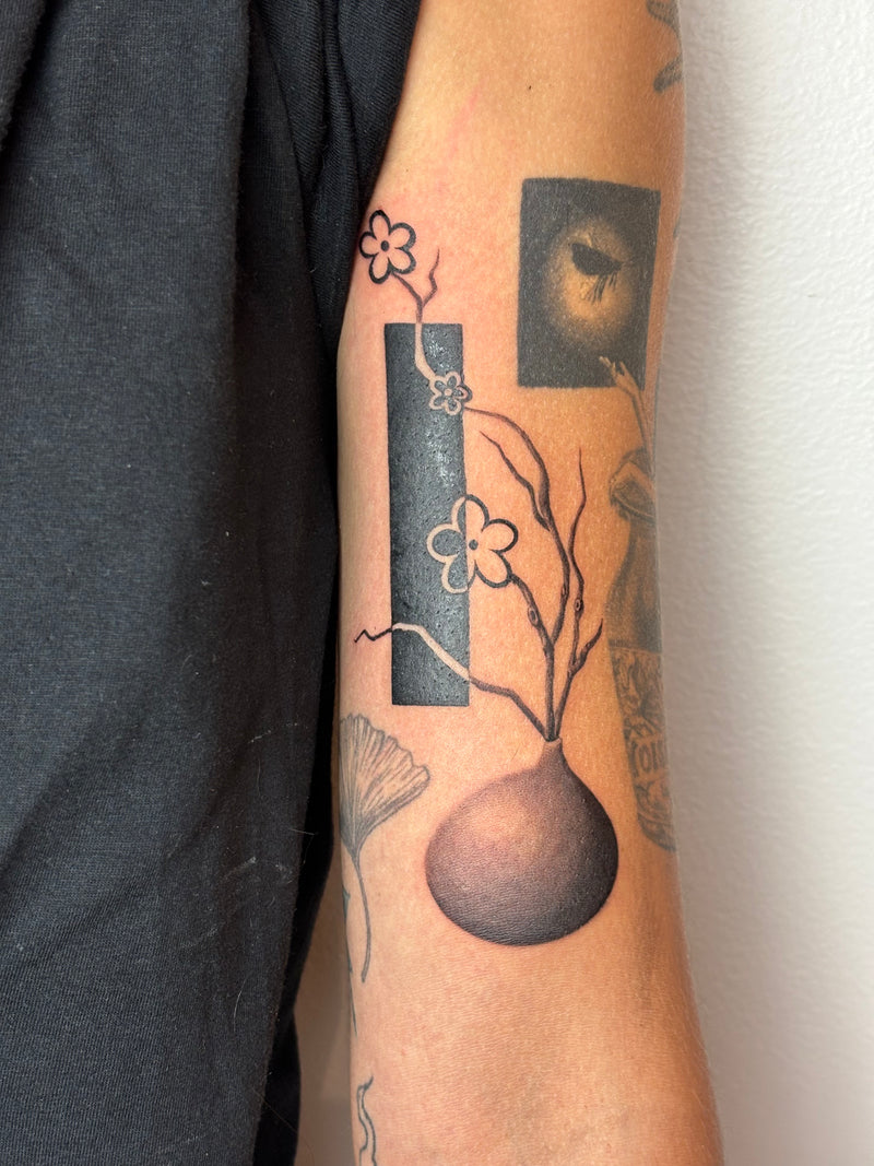 A close-up of a person's forearm featuring a detailed black and gray tattoo design of a vase with flowering branches and geometric shapes.