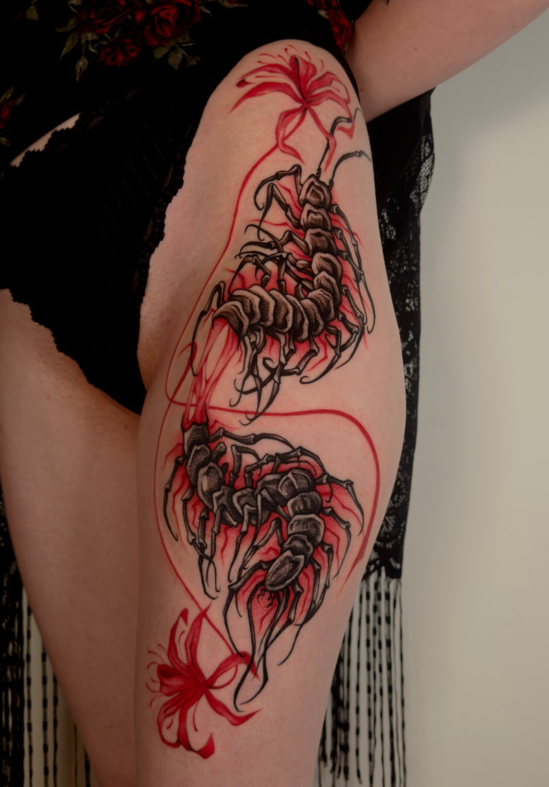 A close-up of a person's upper thigh featuring a detailed tattoo design of two large black scorpions with red and black floral accents, wearing a black lace garment.