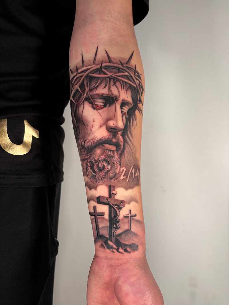 A close-up of a person's forearm featuring a detailed black and gray tattoo of Jesus with a crown of thorns, with three crosses and the date '2/14' below, shown against a plain light gray background.