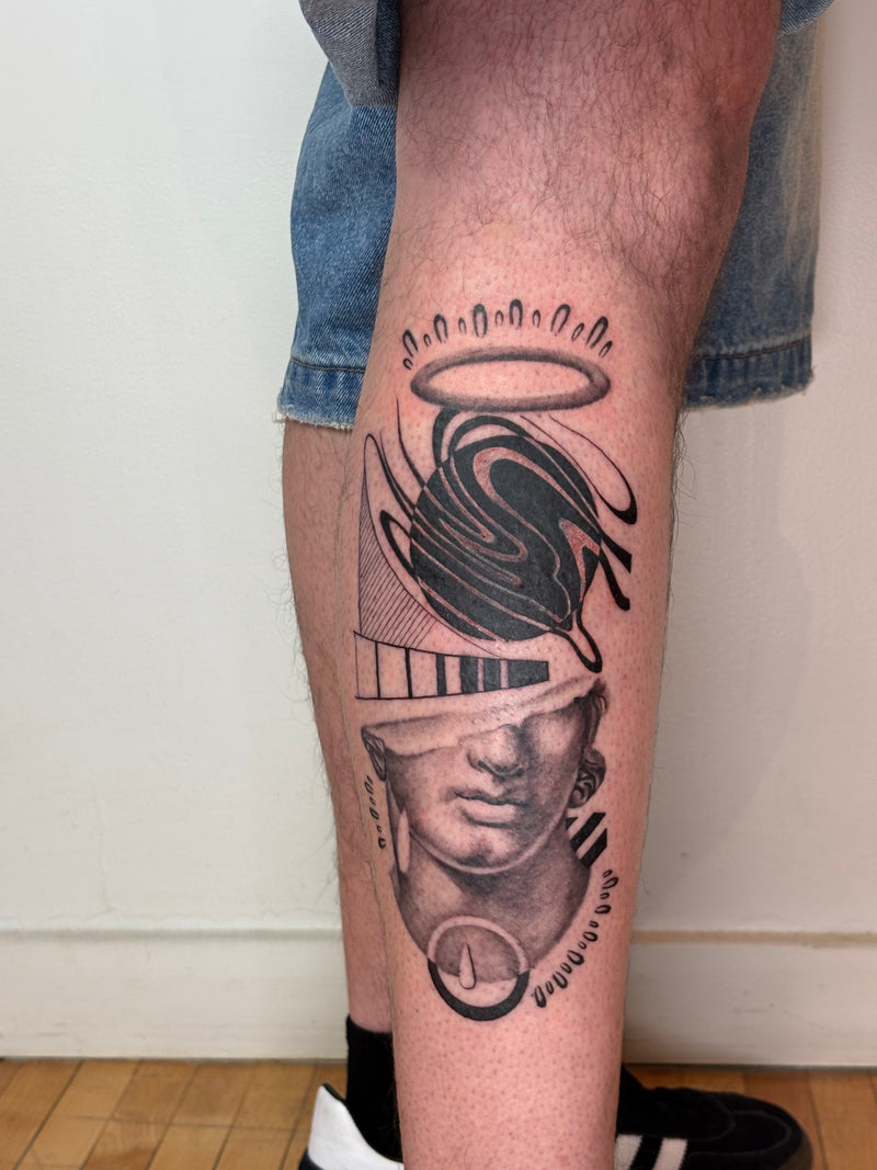 A close-up of a person's lower leg featuring a large black and gray tattoo with a stylized face, geometric shapes, a halo, and abstract flowing lines. The person is wearing denim shorts, black socks, and black-and-white sneakers, standing indoors against a plain light wall and wood floor.