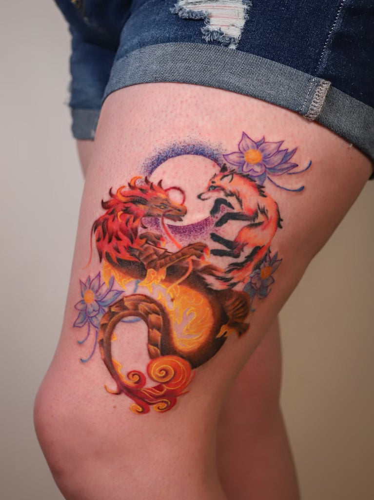 A close-up of a thigh showing a colorful tattoo of a fiery red and orange dragon fighting a fox, surrounded by purple flowers, with a person wearing denim shorts visible at the top.