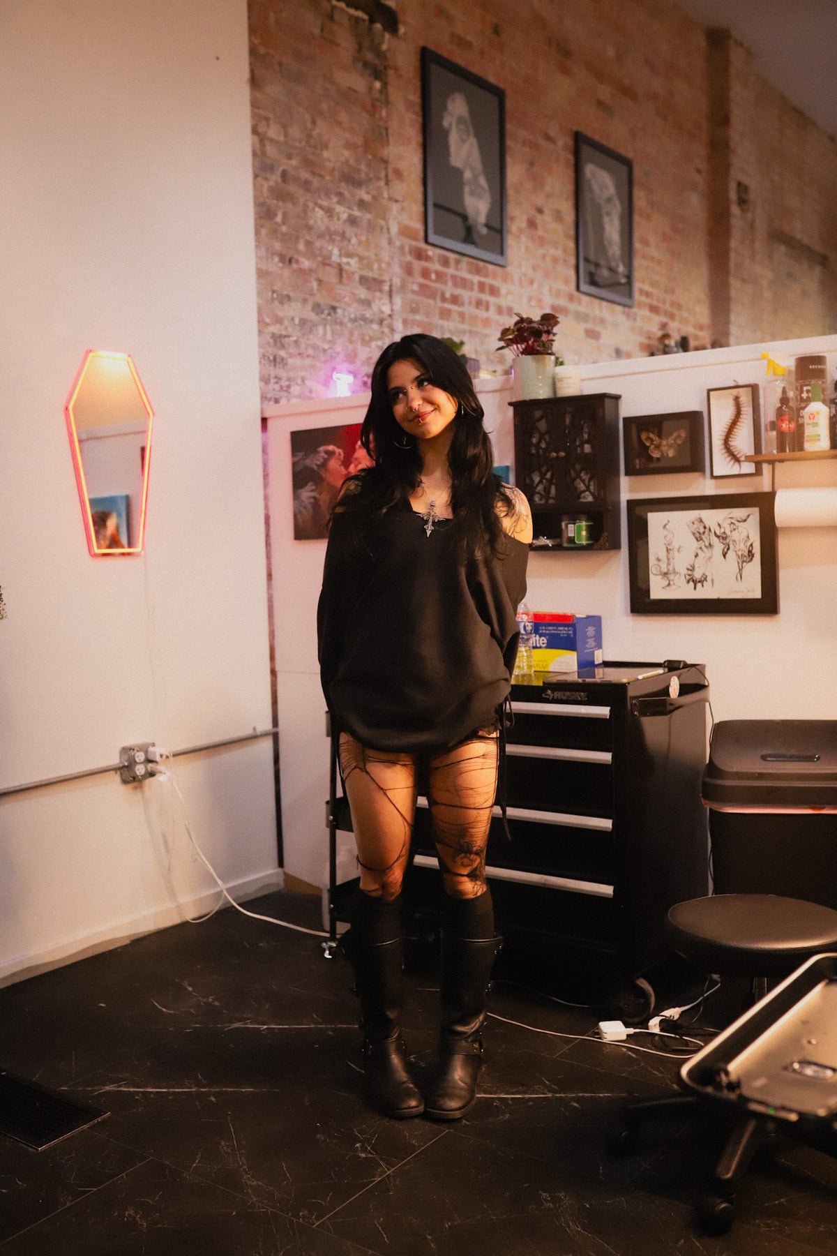 A woman with dark hair stands in a tattoo studio wearing a black oversized hoodie, black thigh-high lace-up stockings, and black knee-high boots. The background features a brick wall, framed art, a tattoo table, and a coffin-shaped wall mirror.