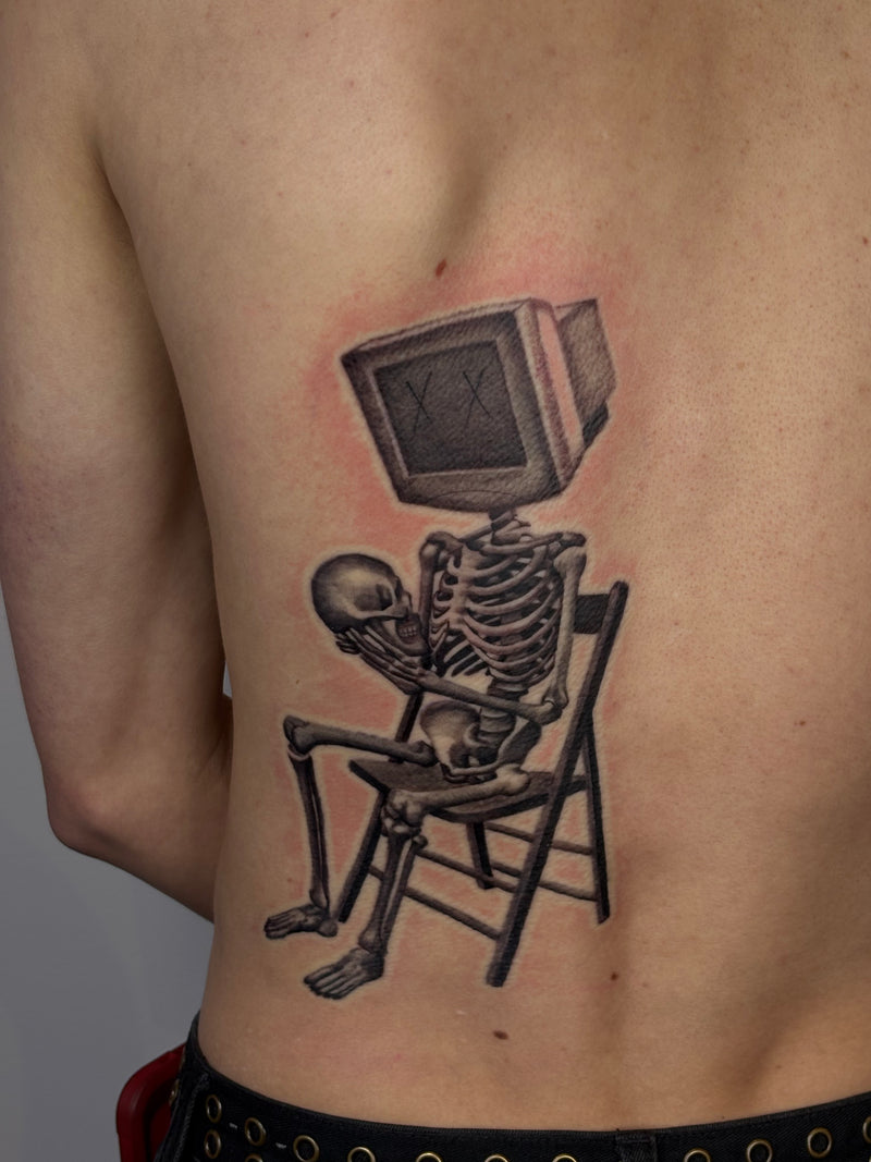 A black and gray tattoo of a skeleton sitting in a chair with a vintage television for a head, located on the back of a person's upper back.
