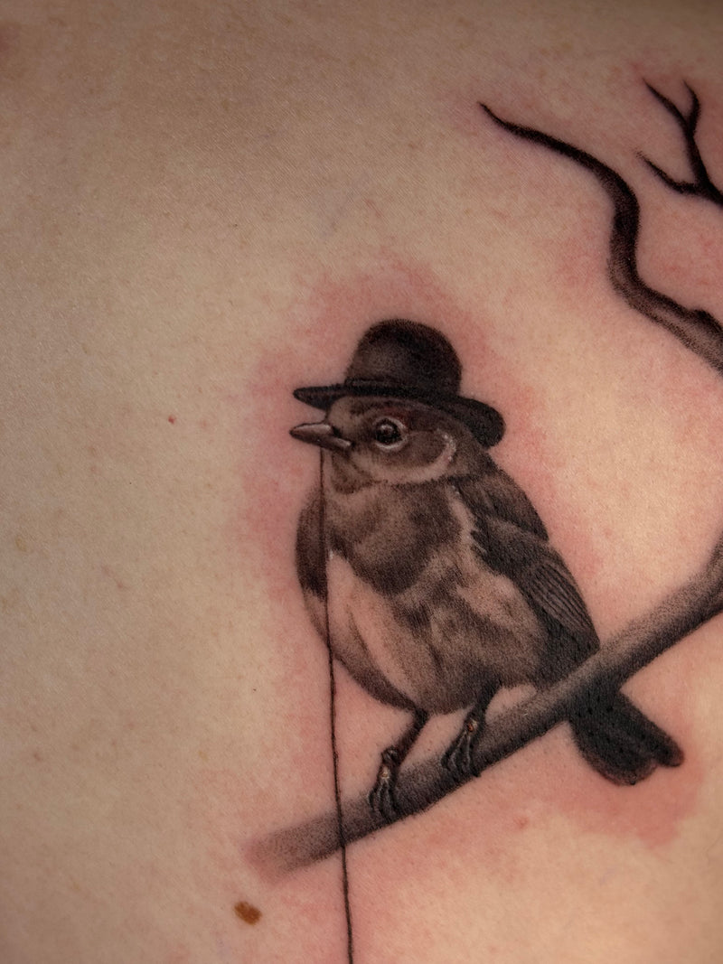 A close-up of a tattoo on skin showing a small bird wearing a hat perched on a branch.