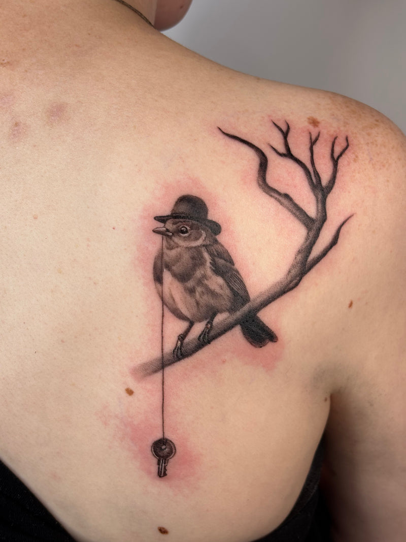 A close-up of a person's upper back showing a detailed tattoo of a small bird wearing a hat perched on a bare branch with a hanging key, surrounded by natural skin tones.
