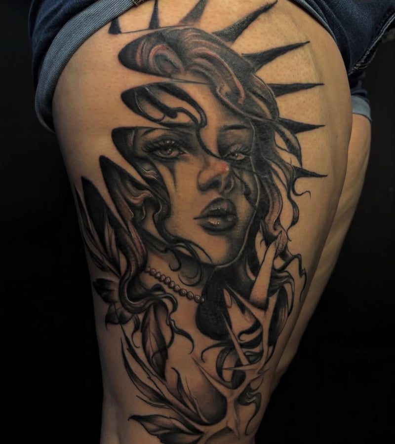 A close-up of a person's upper thigh featuring a large black and gray tattoo of a stylized woman's face with flowing hair and sharp spikes, wearing denim shorts against a dark background.