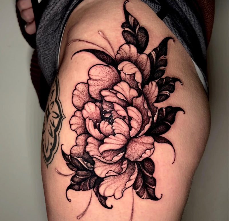A close-up of a large floral tattoo on a person's upper thigh, featuring a detailed black and gray peony design with soft pink shading, shown against skin and casual clothing.
