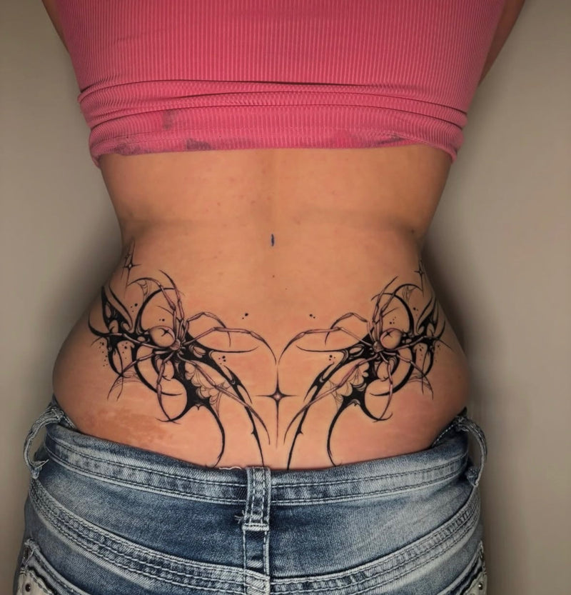A person shown from the back wearing a pink ribbed crop top and blue denim shorts, with a large black tribal-style floral tattoo covering the lower back and upper hips.