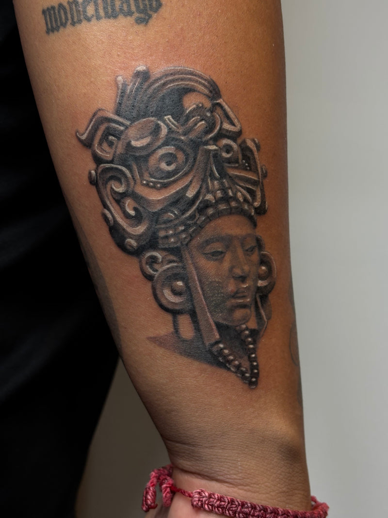 A close-up of a person's forearm featuring a detailed black and gray tattoo of an Aztec-style warrior wearing an ornate headdress. The tattoo is shown on skin with a pink braided bracelet visible at the wrist and a gothic-style tattoo at the upper arm.