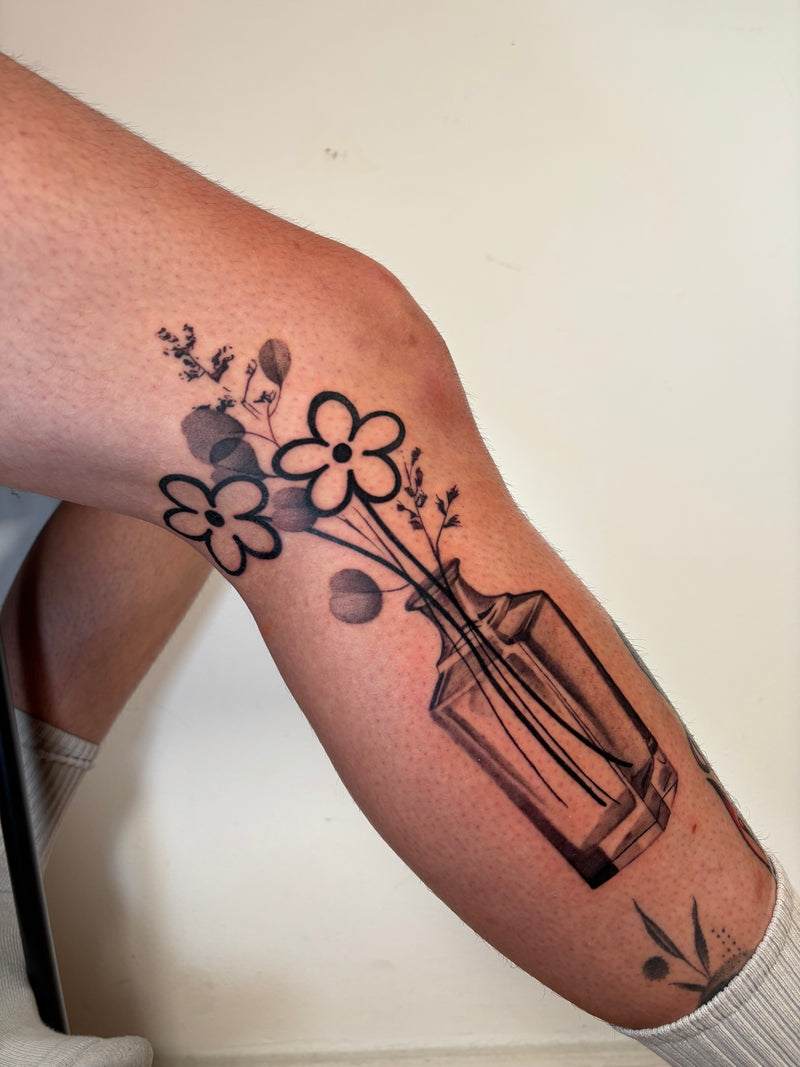 A close-up of a person's lower leg featuring a black ink tattoo of flowers and leaves arranged in a glass vase, shown against a plain off-white background.