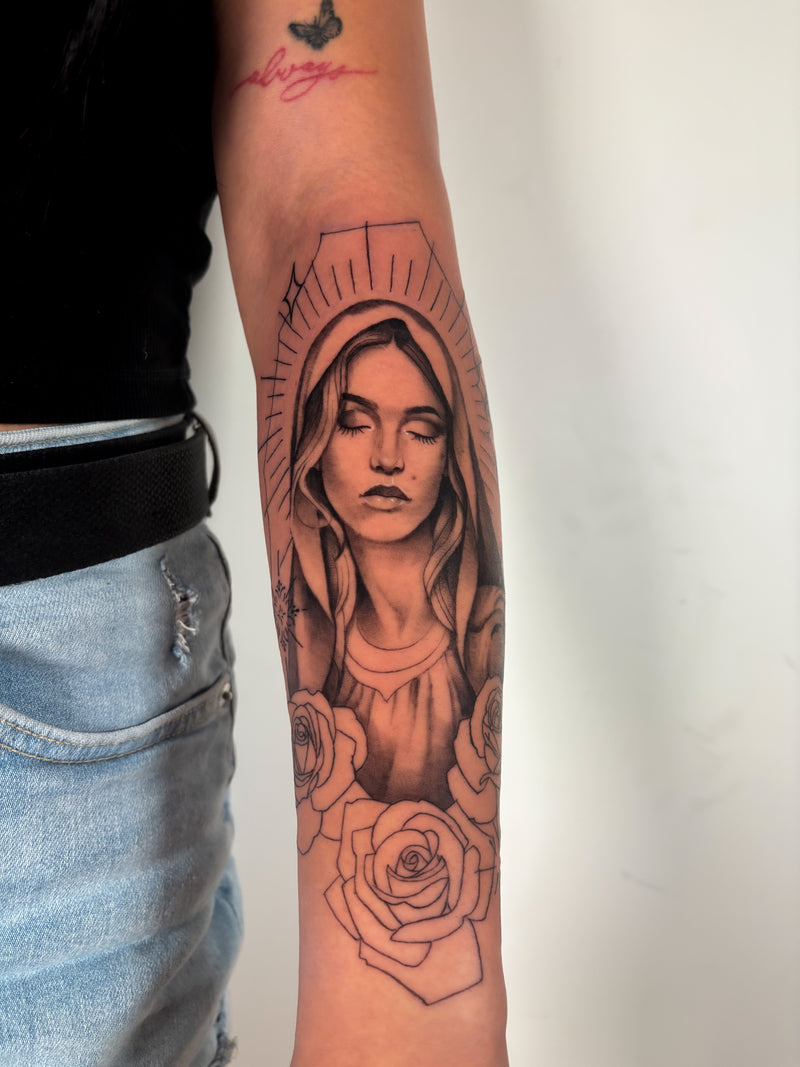 A close-up of a person's forearm featuring a large black and gray tattoo of the Virgin Mary with roses, styled with a black top and light blue jeans against a plain white wall.