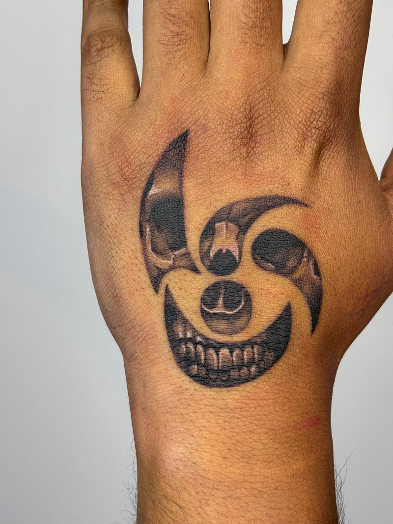 A close-up of a hand showing a freshly tattooed black and gray circular skull design on the back of the hand, set against a plain light gray background.