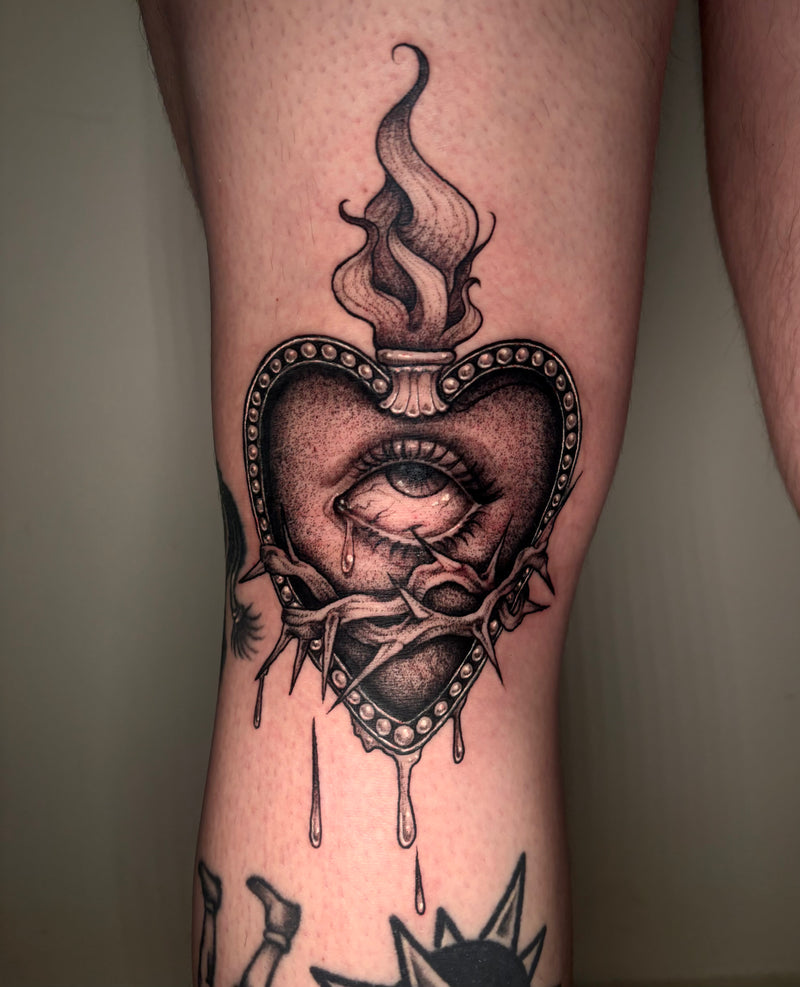A close-up of a leg featuring a detailed black and gray tattoo of a heart with an eye, thorns, dripping details, and a flame rising from the top.