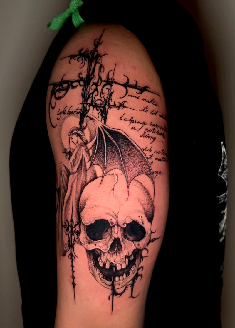 A large black and gray gothic tattoo on a person's upper arm, featuring a skull, a winged figure, barbed wire, and handwritten script text, shown against dark clothing.