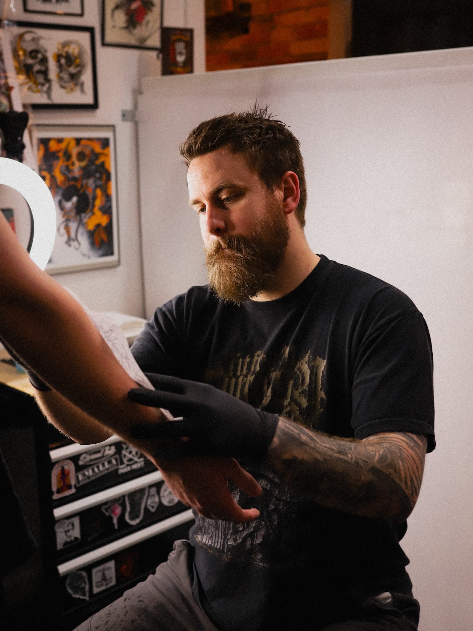 A bearded man with tattoos wearing a black graphic t-shirt and black gloves, sitting in a tattoo studio and applying ink to a client's arm.