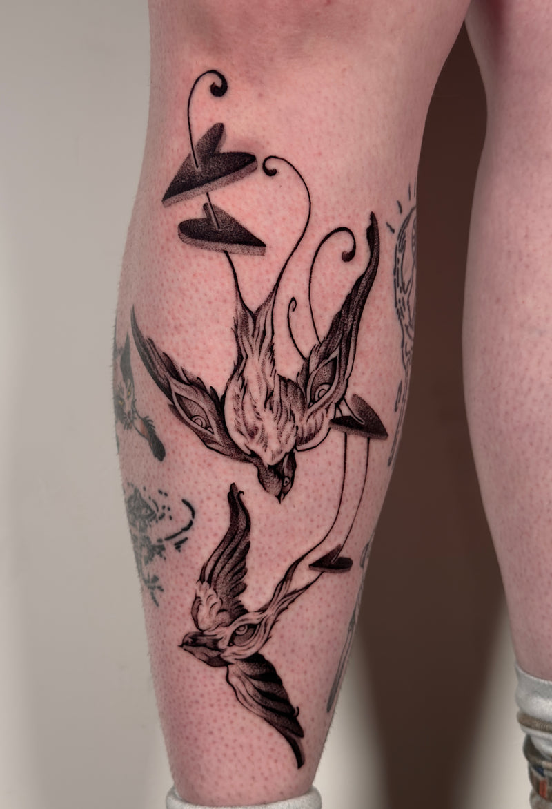 A close-up of a person's calf with a detailed black and gray tattoo of two stylized birds and abstract curved lines, showing the skin texture and surrounding leg area.