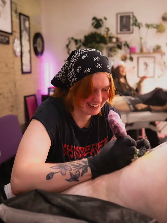 A tattoo artist wearing a black bandana, black t-shirt with red text, and black gloves smiles while tattooing a client's leg in a tattoo studio.