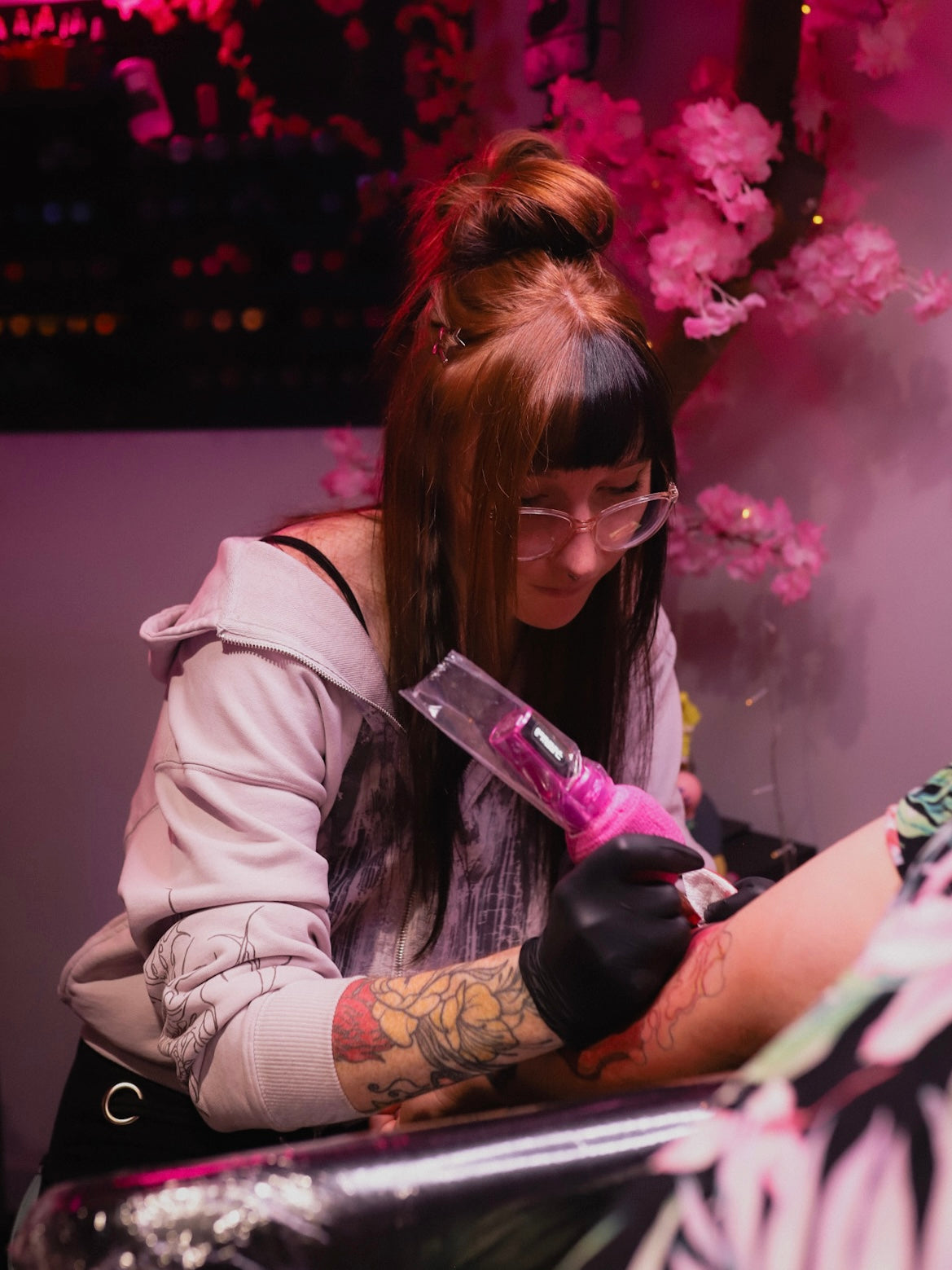 A tattoo artist wearing glasses, a light pink hoodie, and a black glove applies a tattoo to a client's arm using a pink machine in a dimly lit studio decorated with pink cherry blossoms.