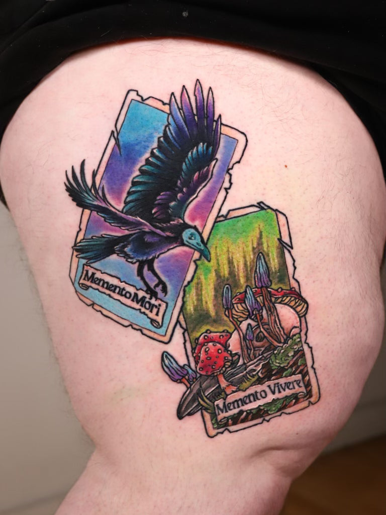 A close-up of a thigh showing a colorful tattoo design composed of two tilted tarot-style cards. The left card features a black and blue raven over a purple-blue gradient background with the text 'Memento Mori'.