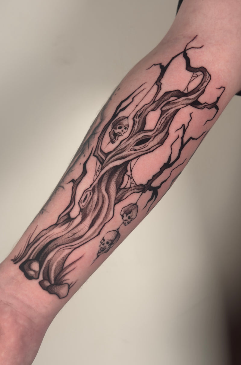 A black and gray gothic-style tattoo of a gnarled tree with skulls on a person's forearm, shown against a plain light background.