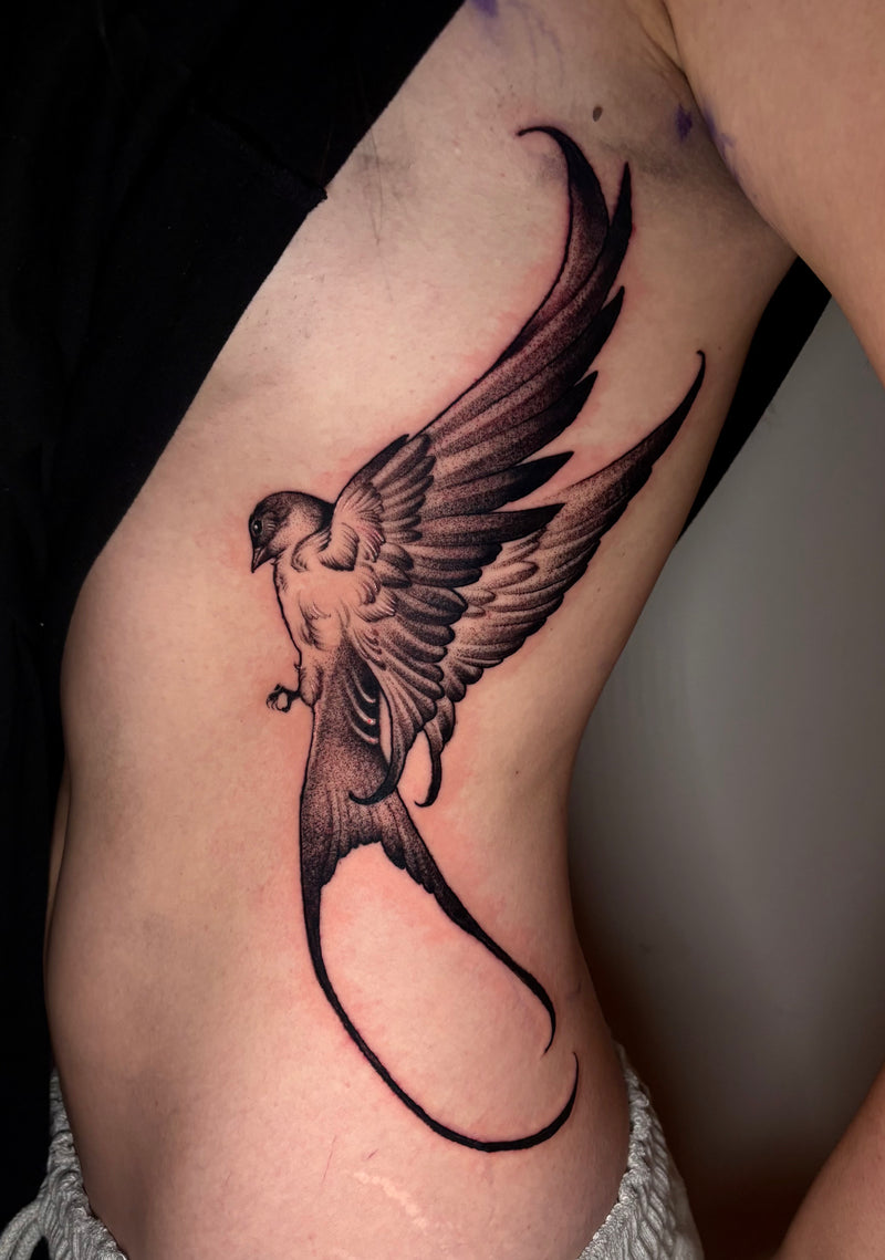 A black and gray tattoo of a stylized bird with spread wings on a person's upper arm, shown in a close-up photo.