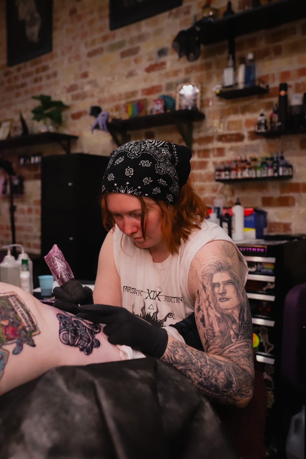 A tattoo artist wearing a black bandana and white graphic t-shirt applies ink to a client's leg in a tattoo studio with a brick wall background.