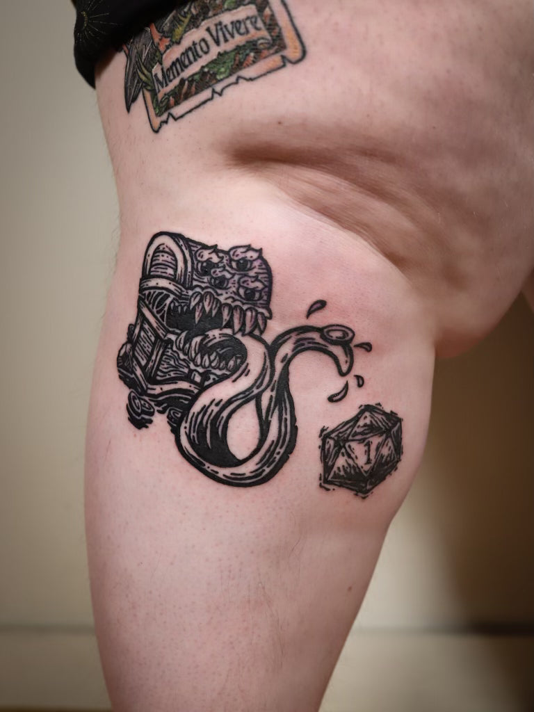 A close-up of a person's upper thigh showing a black ink tattoo of a treasure chest and a die, with another tattoo partially visible near the top.