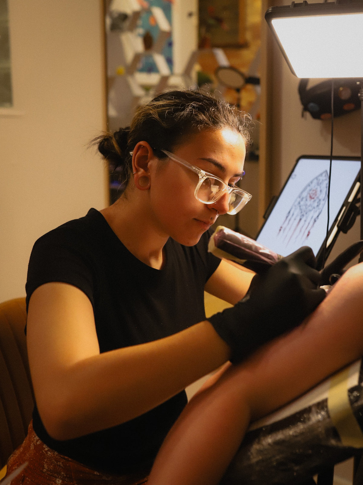 A tattoo artist wearing a black t-shirt and black gloves applies a tattoo to a client's leg using a tattoo machine and a light box in a studio setting.
