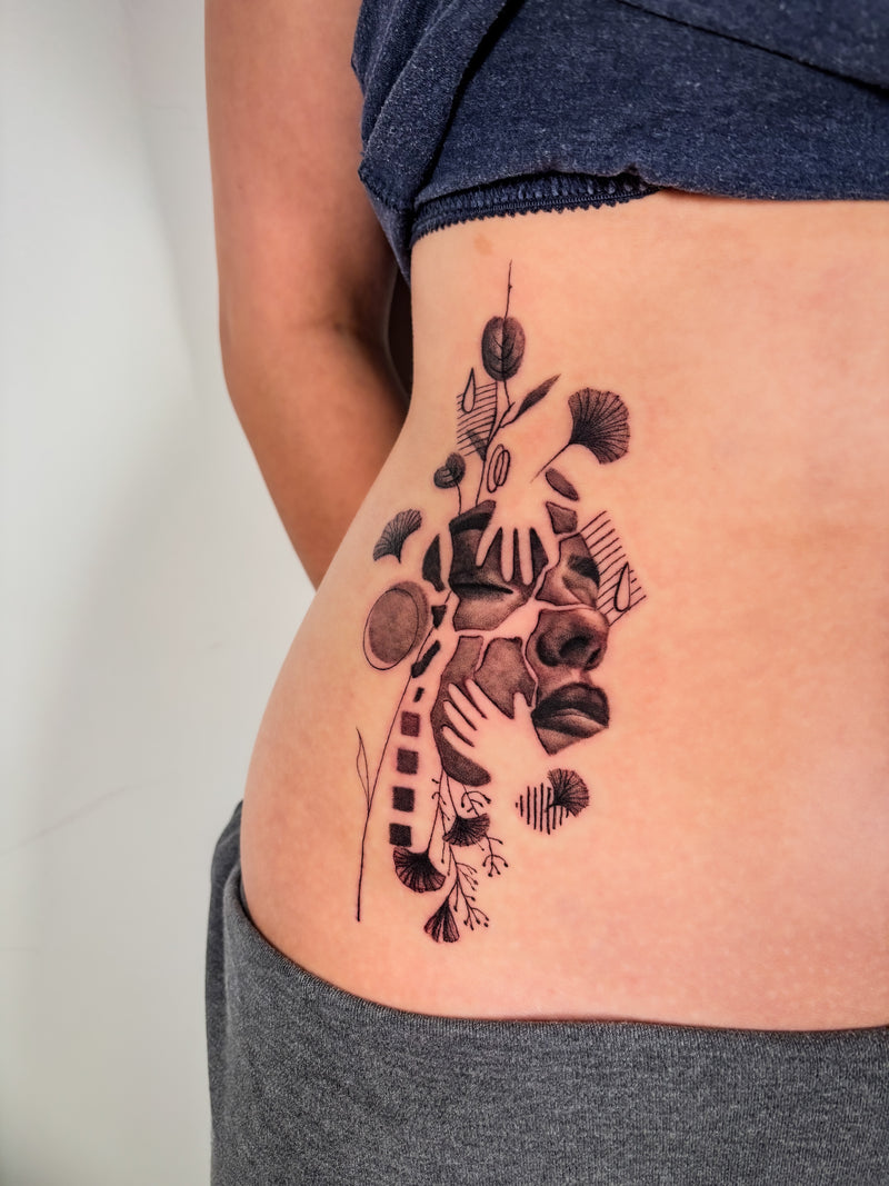 A close-up of a person's side torso showing a black and gray floral and abstract tattoo design on the hip, wearing a dark blue top and gray shorts.