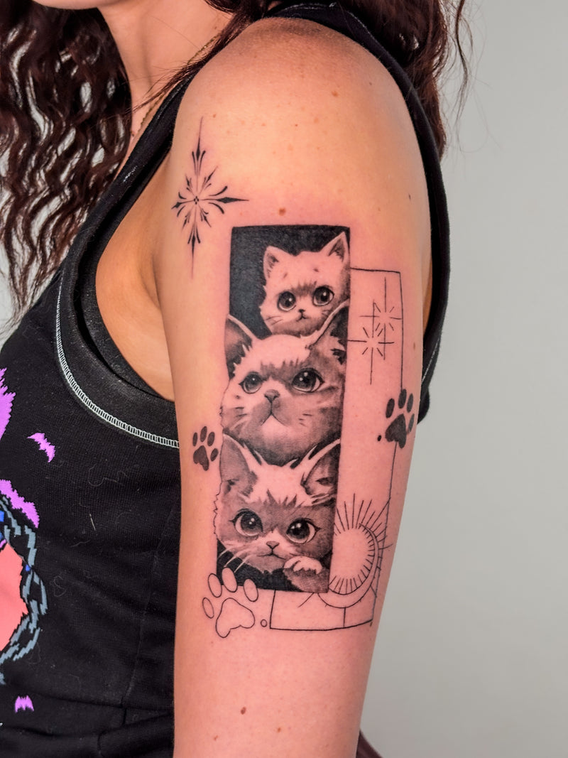 A close-up of a person's upper arm and shoulder showing a black and gray tattoo of three cats stacked vertically with paw prints and geometric star details.