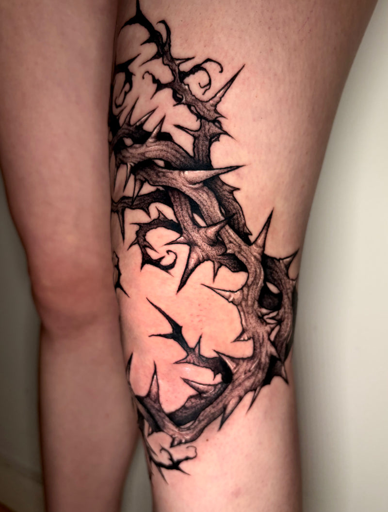 A close-up of a person's leg featuring a detailed black and gray tattoo of thorny branches wrapping around the skin.