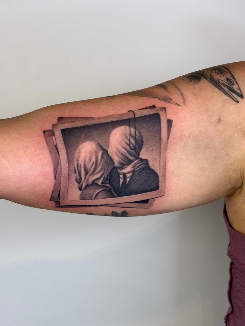 A close-up of a person's upper arm featuring a realistic black and gray tattoo of two people in hoodies, with a small stack of tilted photo frames behind them.
